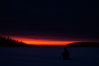 Orange dusk band over a frozen lake with gear silhouettes near a hole